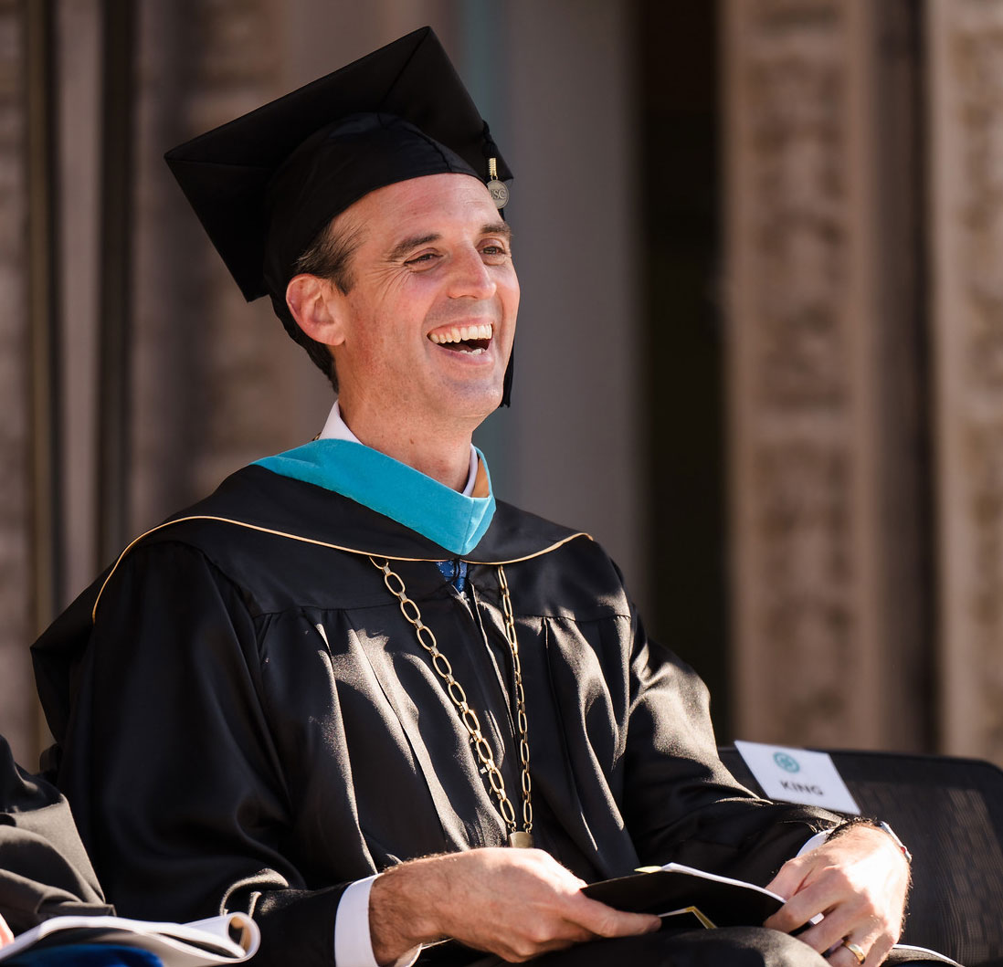Greg Smith at Commencement Greg Smith wears a black graduation cap and gown with a teal sash and a chancellor medallion