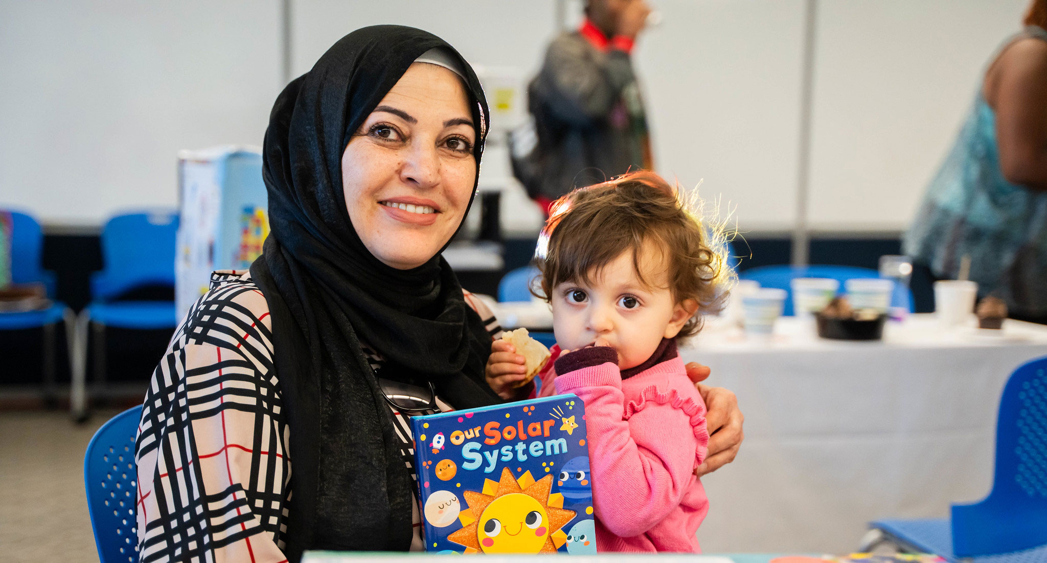 Childcare center A woman sitting down at a table is holding a small child and a children's book