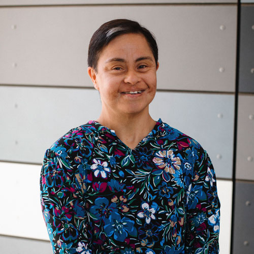 SDCCD student Ivette, is smiling in front of a gray and white paneled wall, wearing a colorful floral blouse.