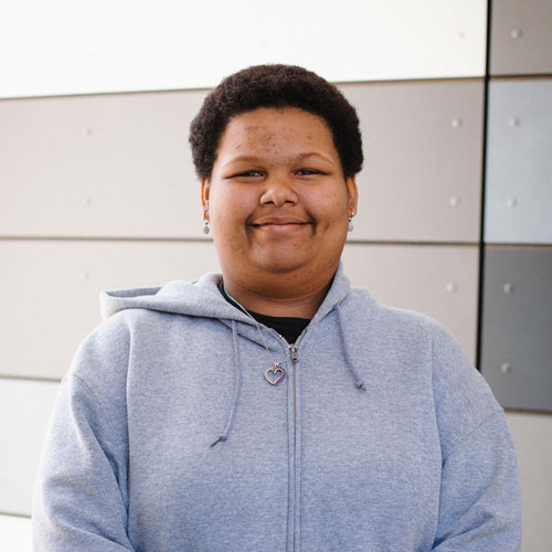 SDCCD Student Kamille is smiling, wearing a light gray hoodie with a gray patterned wall in the background.