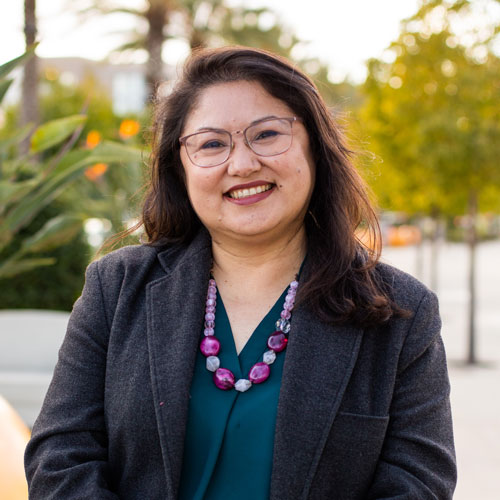 Lilian Garcia is smiling, she is wearing a green shirt and gray blazer with a chunky necklace in pink shades.