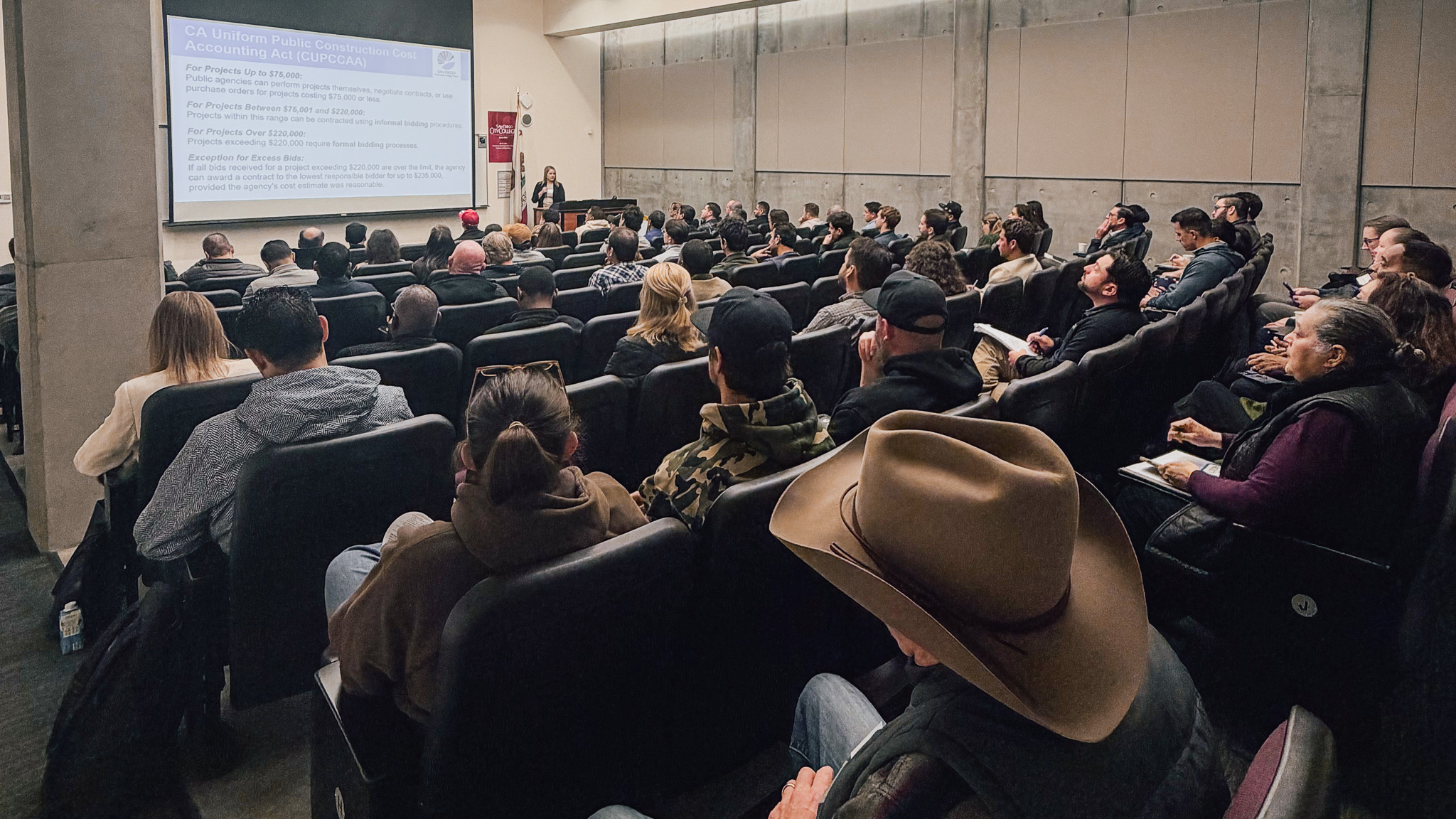 A large room with slight stadium seating almost completely filled with men and women seated and watching a presentation happening at the front of the room, which includes one female talking and a project showing a rendering of the building project being discussed.