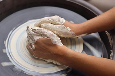 Two hands covered in clay working on a pottery wheel with a lump of clay being pressed and spun on the wheel.