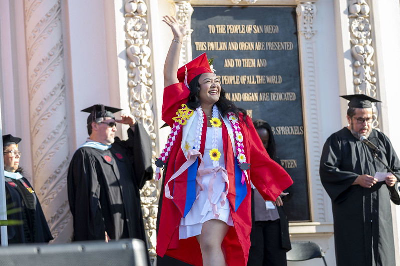 
A student in a red cap and gown waves to the audience as she walks across the stage to receive her degree.
