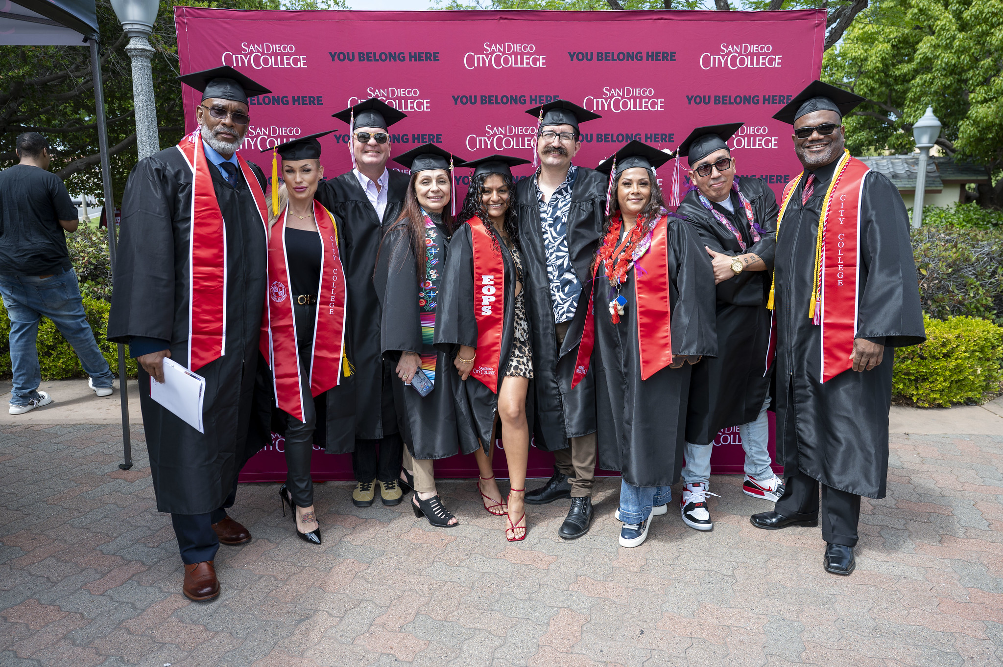 
Nine students in black graduation caps and gowns with a red sash in front of a red background with the City College logo.
