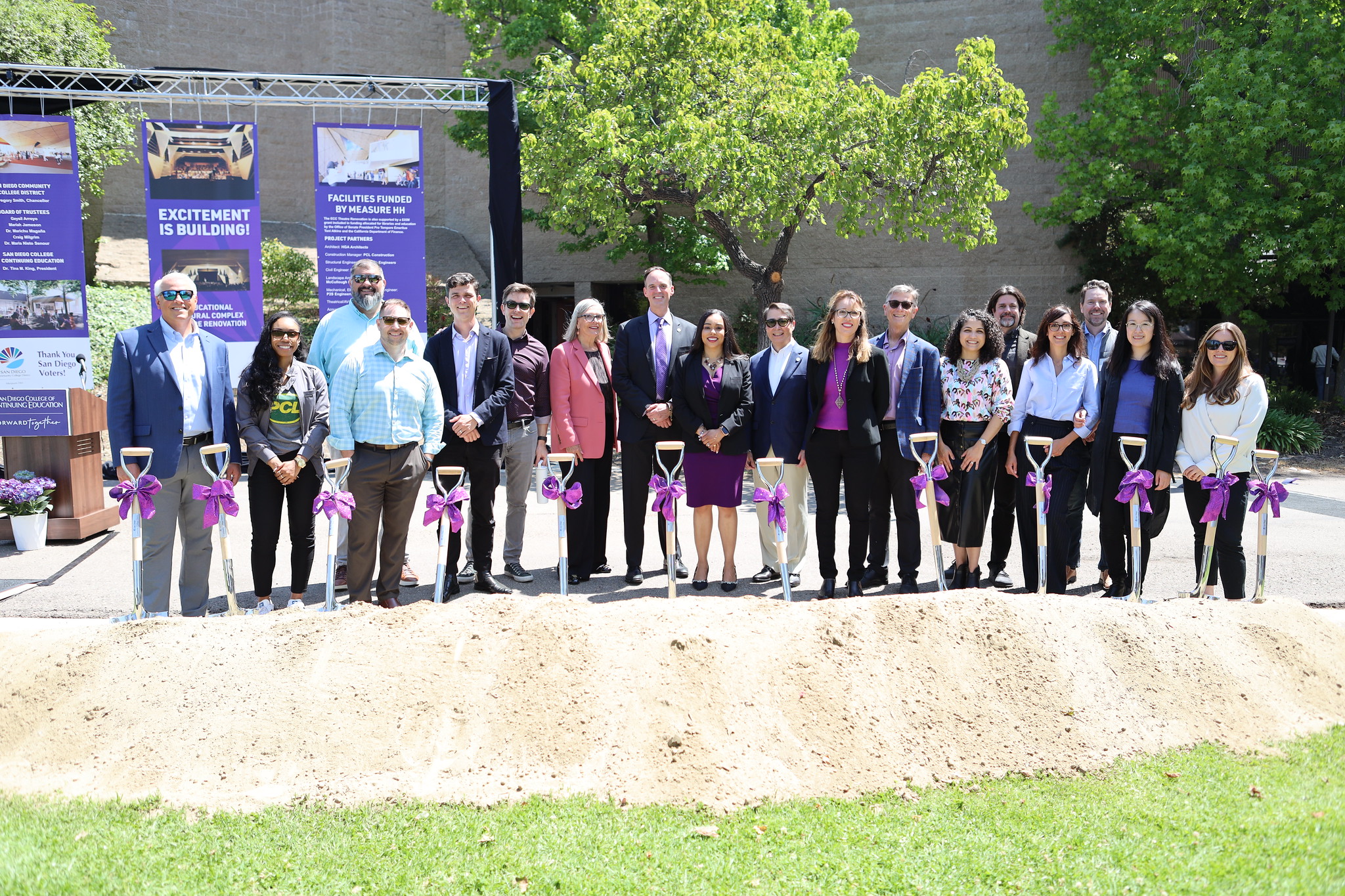 
18 people stand behind a pile of dirt and shovels with bows on them.
