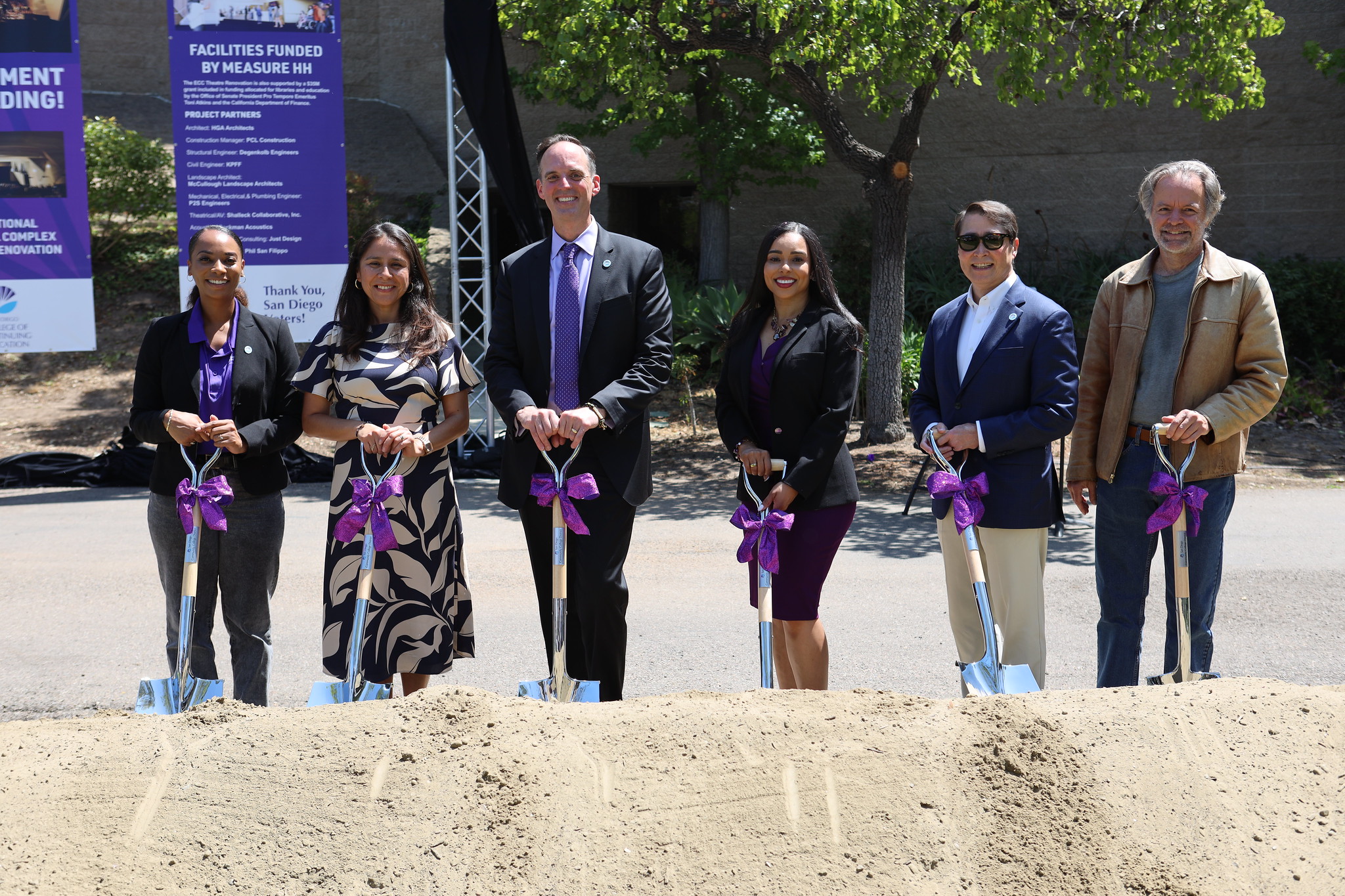 
Six people stand behind a pile of dirt holding shovels with large purple bows on them.
