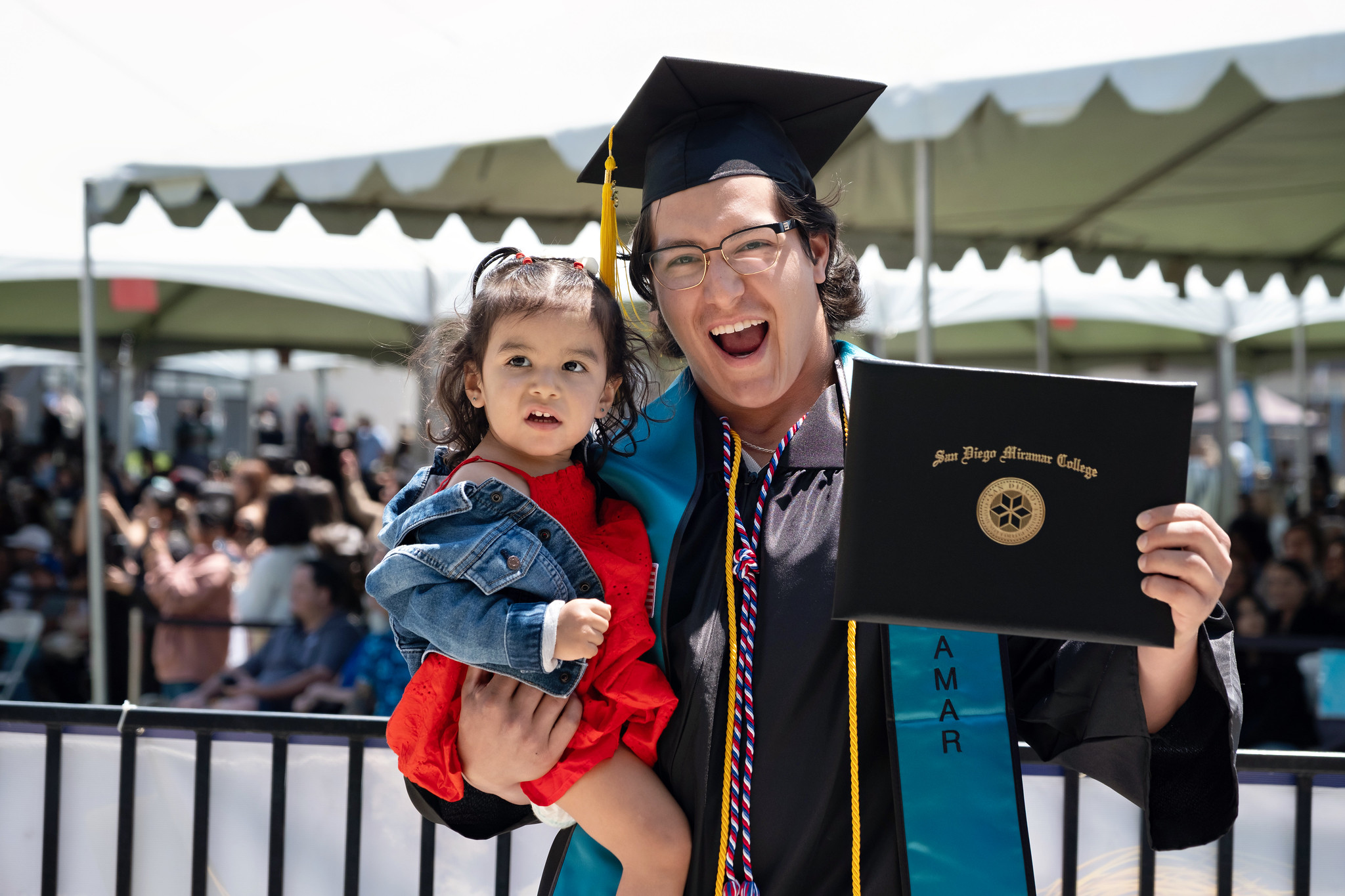 
A smiling graduate is holding his toddler daughter in one hand and his degree with the other.
