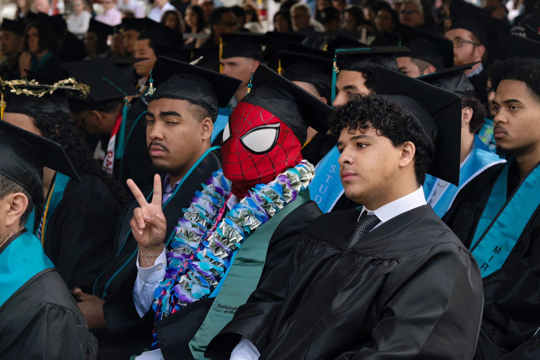 
A group of graduates, including one in a Spider Man mask, are seated at commencement.
