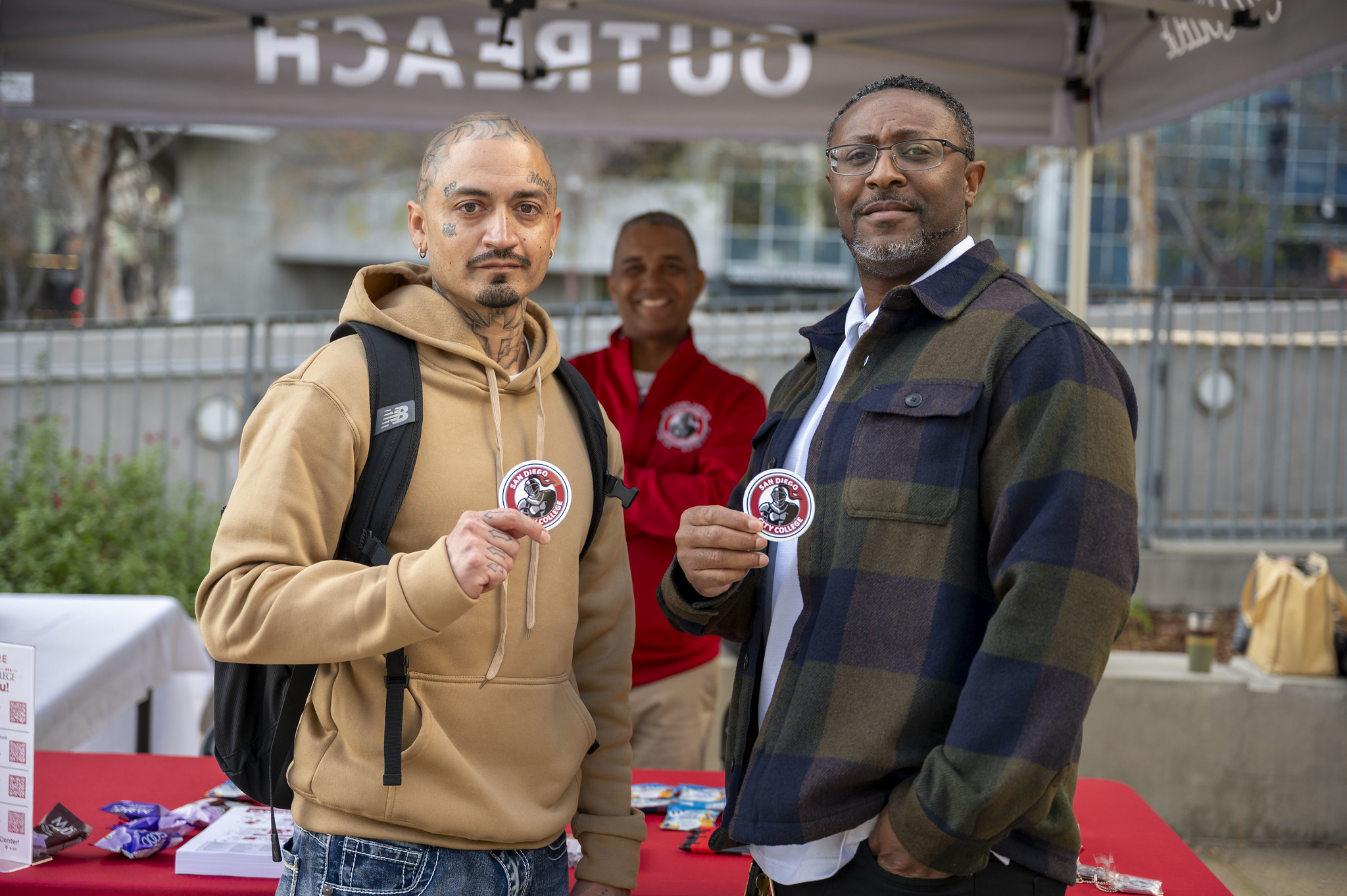 
Two men hold up City College stickers. City College President Ricky Shabazz is behind them at the welcome table.
